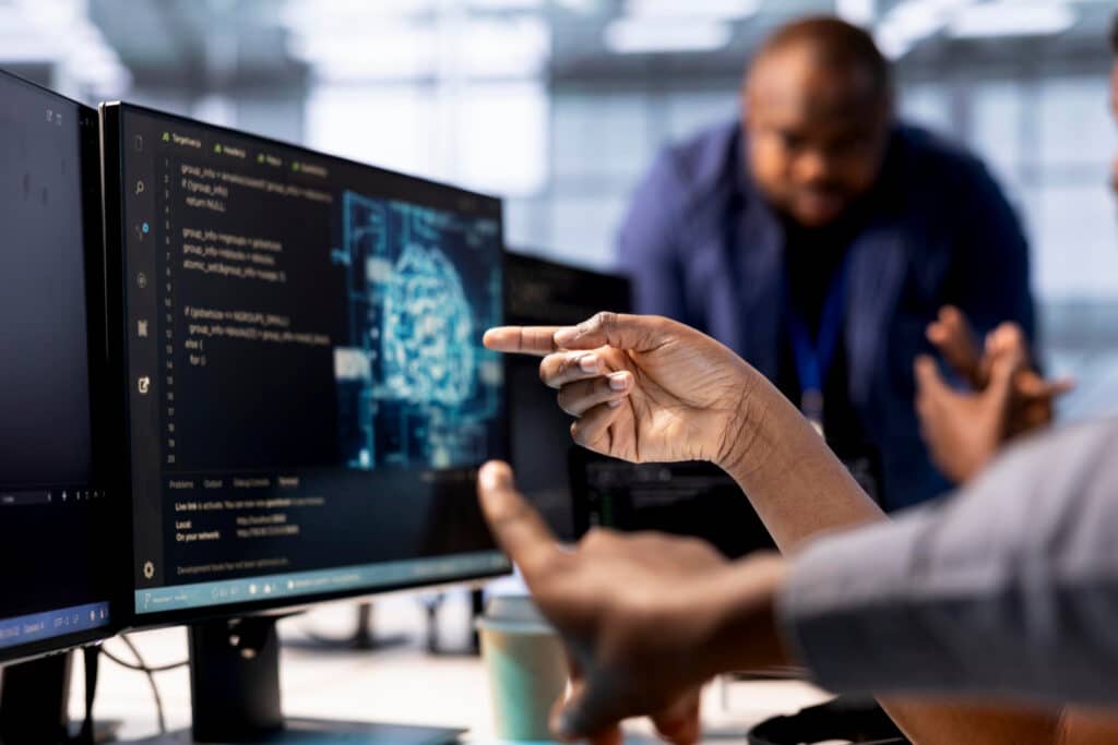 Team members collaborating at a desk, one person pointing at a code editor on a monitor while another watches nearby.